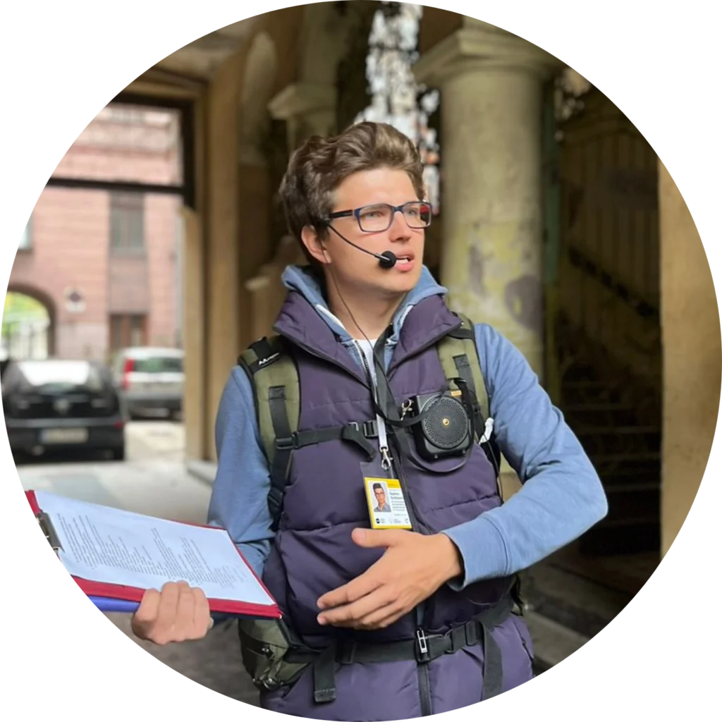 W kole przewodnik patrzący na grupę, niebieski kolor ubrania. W prawej ręce trzyma teczkę, a lewą gestykuluje. Obok ust mikrofon wzmacniacza głosu.